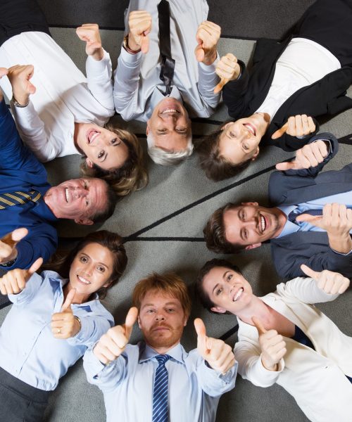 Eight smiling at camera business people lying on floor in circle and showing thumbs up. Top view.